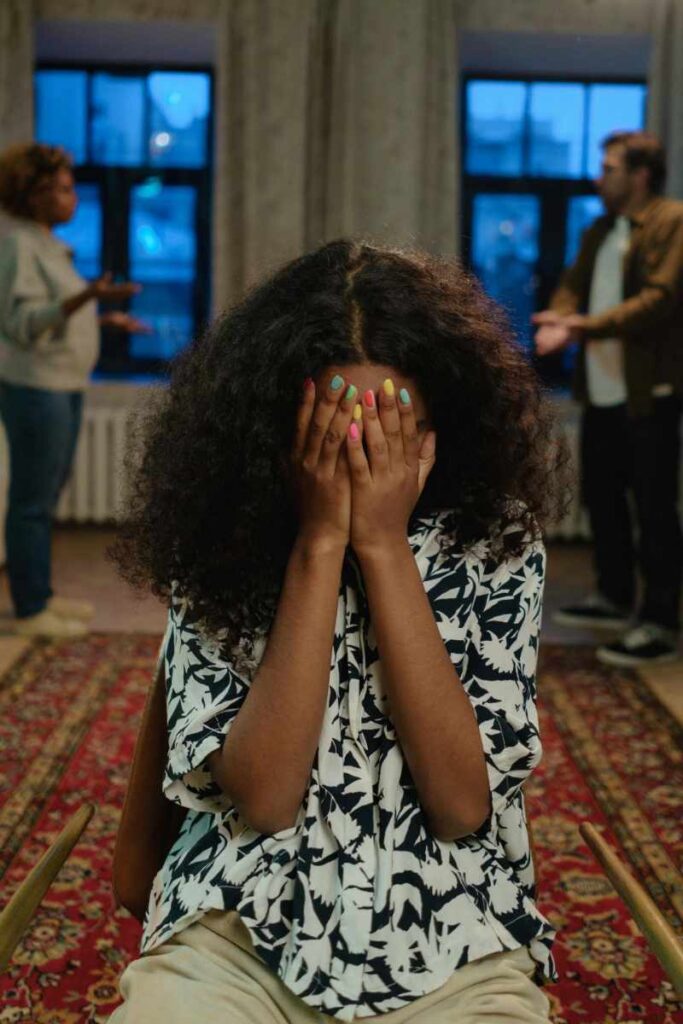 Young girl covering her face in distress while parents argue in the background, symbolizing the emotional impact of family conflict and estrangement.