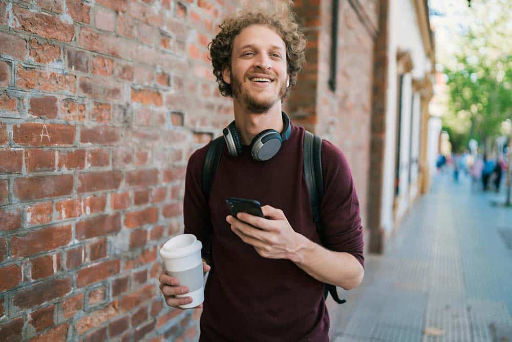 A young man with curly hair and a beard smiling while walking down a city sidewalk.