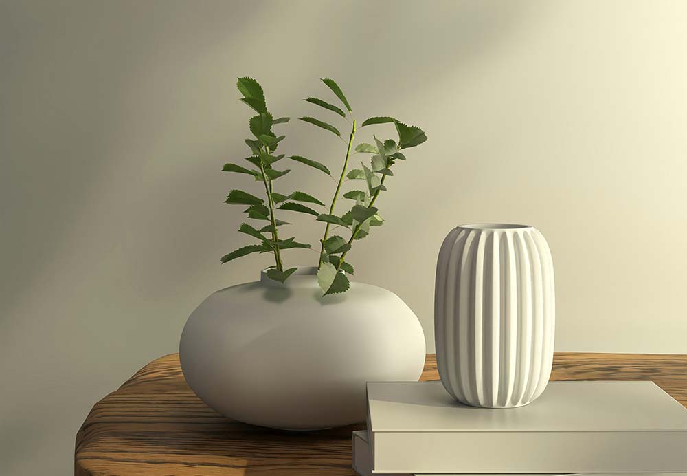 Two white ceramic vases and a stack of books sit on a rustic wooden table against a plain wall with soft leaf shadows.