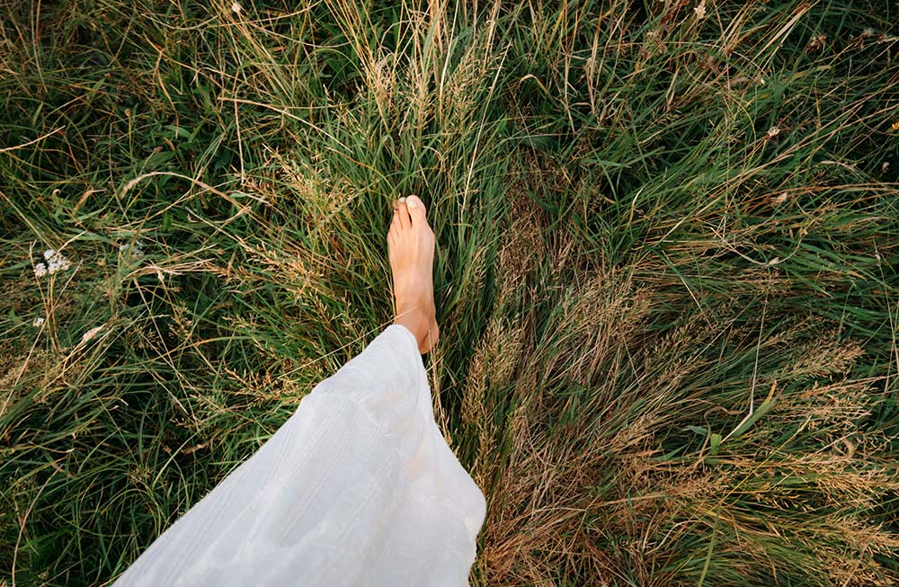 A first-person view of a bare foot stepping through tall, wild green and golden grass, with the edge of a white flowing dress visible.