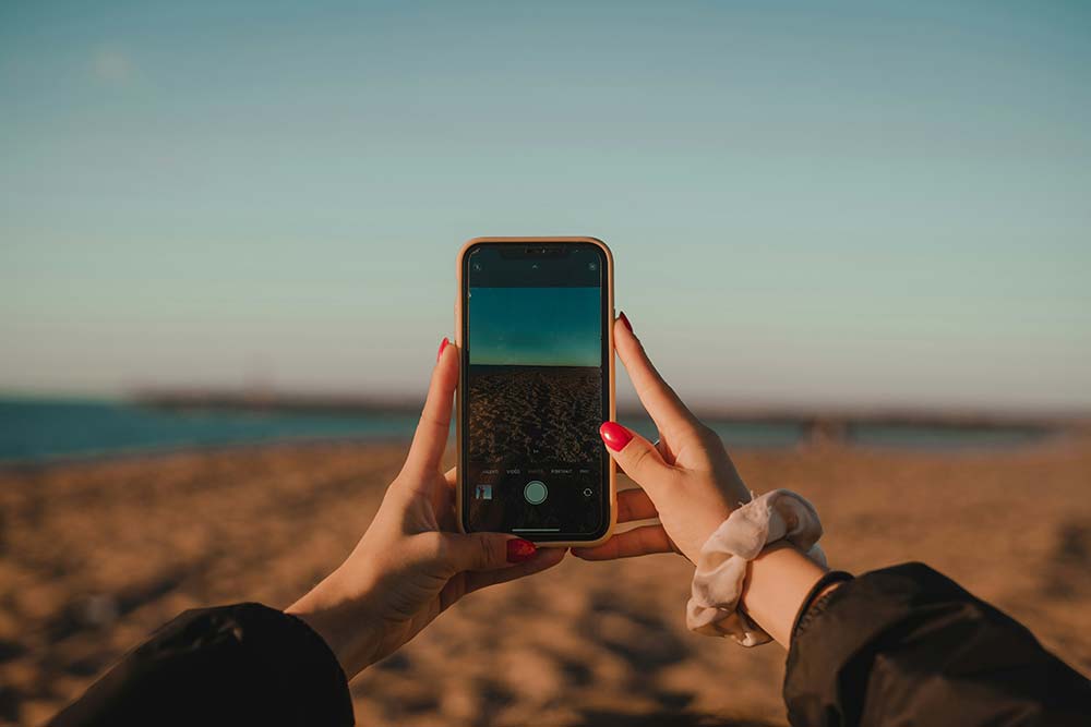 A first-person perspective shows a person’s hands holding a smartphone to take a photo of a beach at sunset.